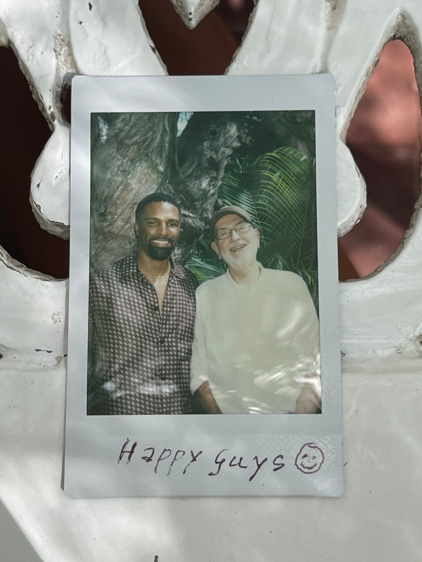 Instant photo of two smiling men standing together in front of a palm tree, handwritten note: happy guys